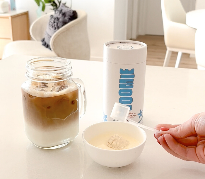 A glass mason jar mug with a layer of milk followed by a layer of coffee. Next to the glass sits a cylinder of Voomie Flow Fuel, and a person's hand is putting a scoop of Flow Fuel powder into a small dish filled with cream.