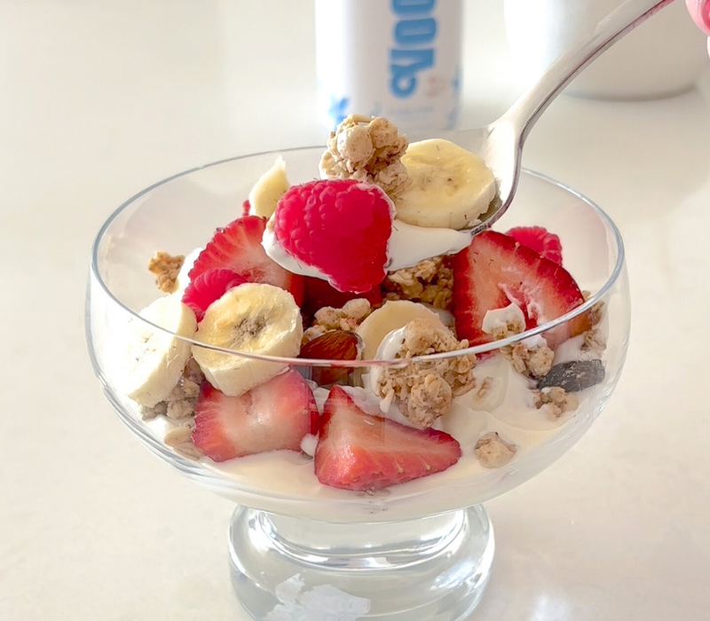 Glass bowl of yogurt topped with banana, strawberries, raspberries, and granola, with a spoonful lifted—ideal morning meal to enjoy alongside Flow Fuel by Voomie.