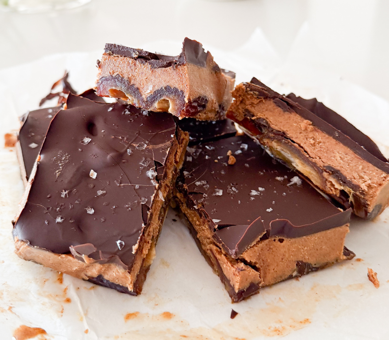 Close up image of stack of almond butter Bedtime Bliss slice squares, with the layers of dates, almond butter, chocolate and flaky sea salt are visible.