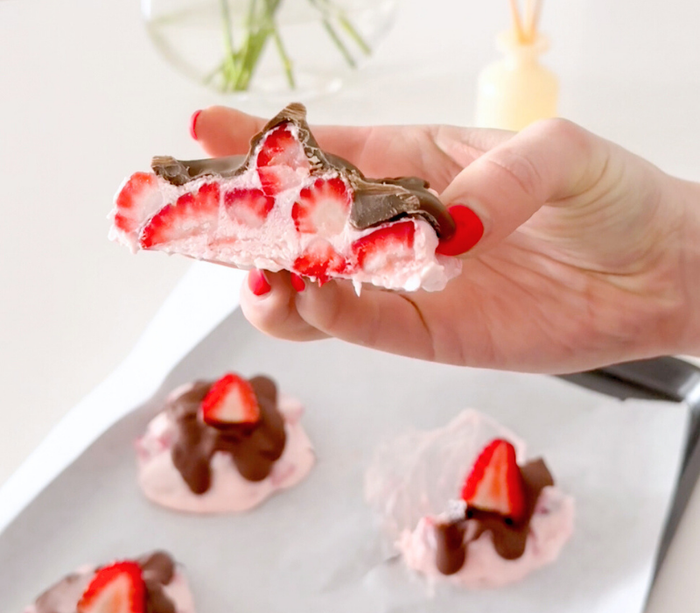 A hand holding a half of a Chocolate Strawberry Yoghurt Bite . You can see the layer of chocolate on top and the strawberries within the yoghurt