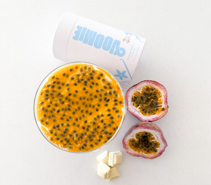 Above shot of overnight oats with a layer of white chocolate and passionfruit on top, with a cylinder of  Voomie Flow Fuel, a halved passionfruit and some white chocolate placed around the bowl.