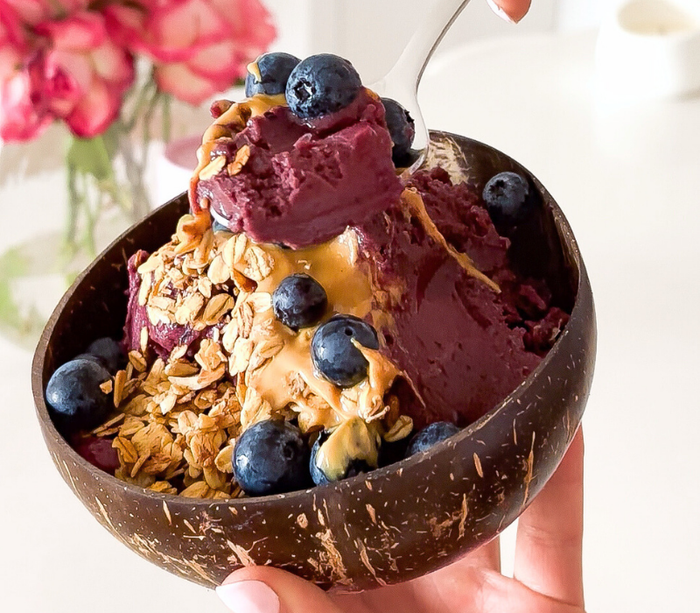 A close-up shot of a spoonful of rich, creamy acai sorbet lifted from a smoothie bowl, topped with fresh blueberries, granola, and peanut butter in a rustic coconut bowl.