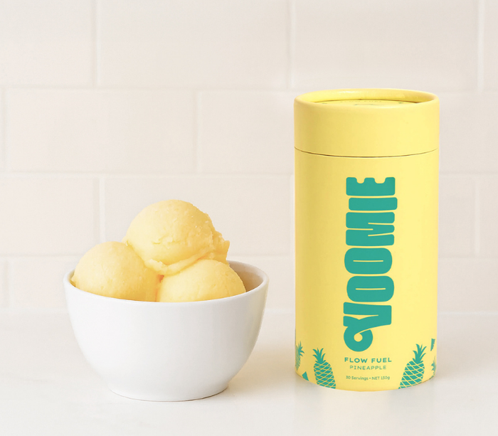 A clean and vibrant image featuring a white bowl of Pineapple Flow Fuel Sorbet alongside a bright yellow Voomie Flow Fuel Pineapple supplement container, set on a white kitchen counter with a tiled backsplash — showcasing a fresh and energising aesthetic.