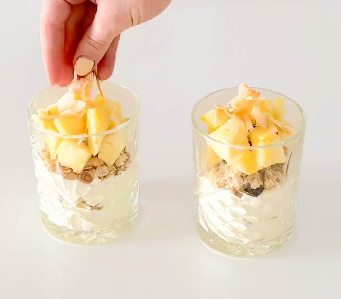A hand placing coconut flakes on top of an assembled pina colada-inspired yoghurt cup layered with yoghurt, muesli, pineapple chunks and coconut flakes.